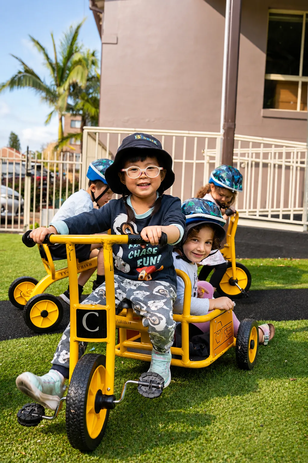 Children riding a yellow trike with a passenger seat across the grass, two more riding behind them